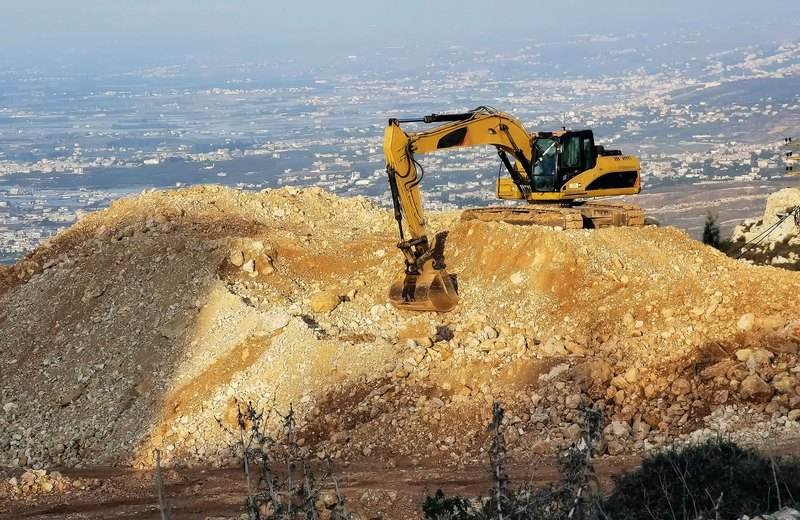 Excavadora amarilla sobre una loma rocosa con la ciudad de fondo