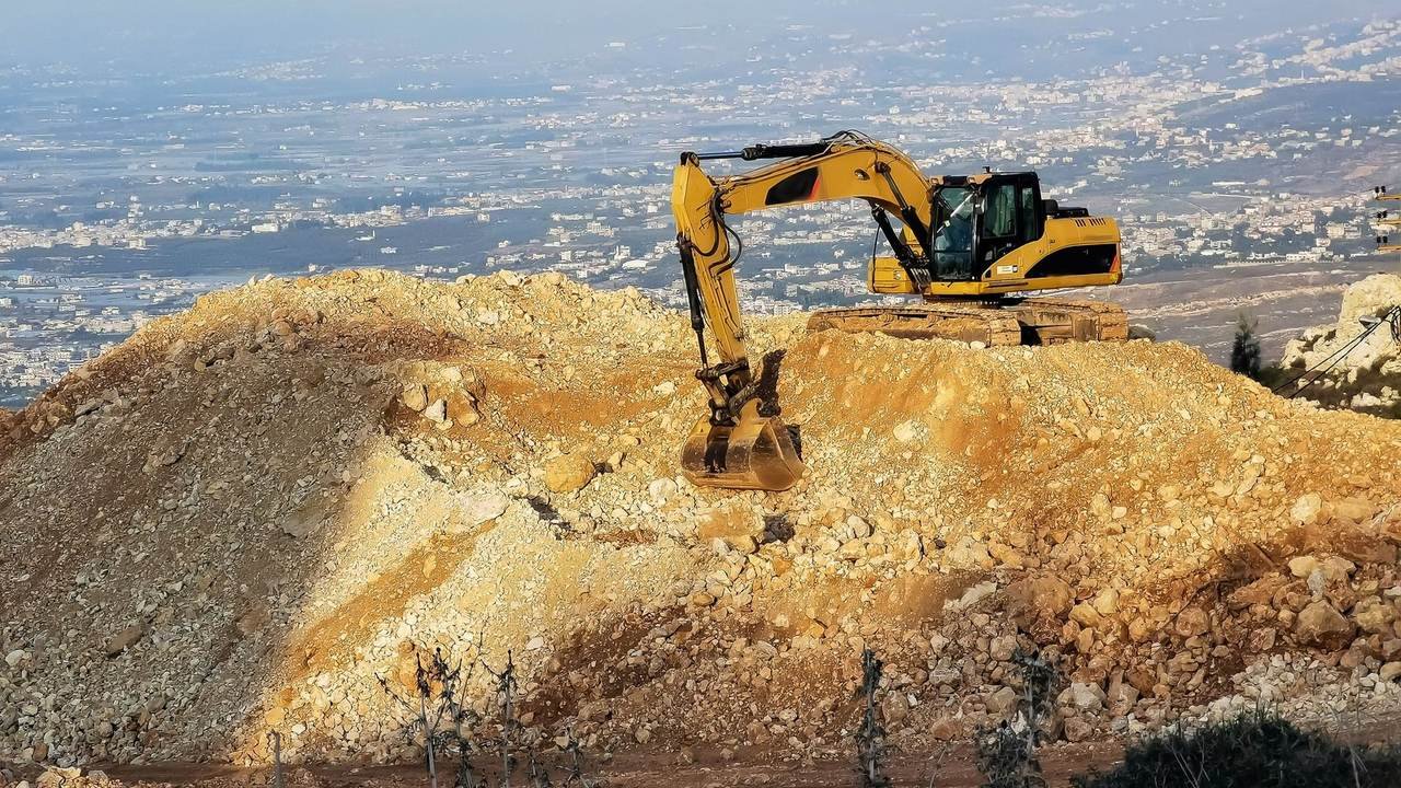 Excavadora amarilla sobre una loma rocosa con la ciudad de fondo