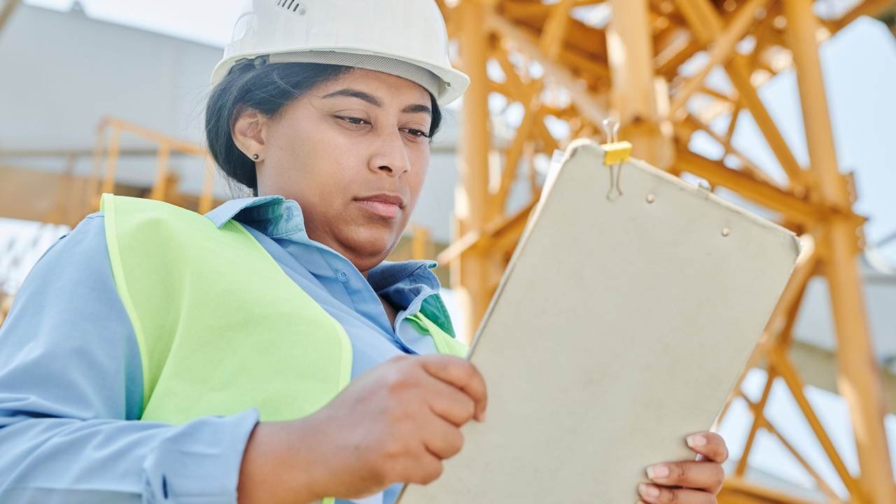Trabajadora con casco inspeccionando una obra con una planilla en la mano